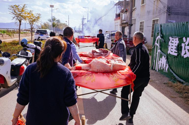 The local family carrying the dowry offering from the pavilion to their house down the street. Notice the smoke in the distance from the firecrackers going off. The lady in the foreground is carrying ducks and chickens in a basket.