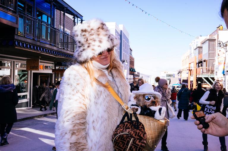 A woman in a spotted fur hat and cream fur coat walks down Main Street carrying a designer bag, festival crowds behind her.