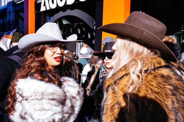 Two women in cowboy hats and fur coats face each other mid-conversation on a crowded Main Street.