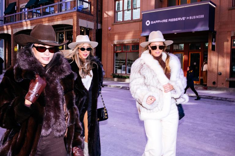 Three women in fur coats, wide-brimmed hats, and sunglasses stride down Main Street past a Chase Sapphire Reserve sign.