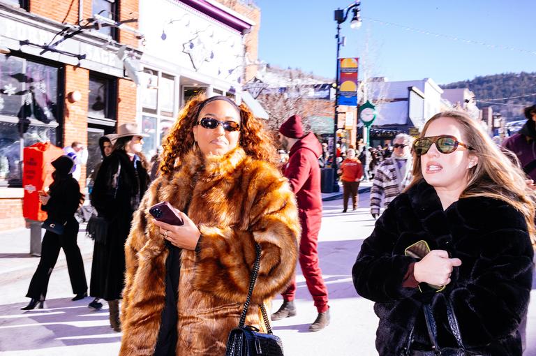 Two women in fur coats walk down Main Street in bright afternoon light, a figure in a red onesie visible behind them.
