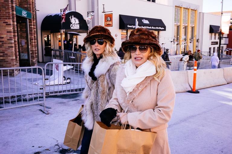 Two women in fur-trimmed hats and coats carry shopping bags outside a storefront on Main Street.