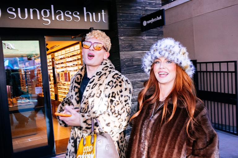 Two people laughing outside the Sunglass Hut, one in a leopard print coat with orange-tinted sunglasses and the other in a fur hat and brown fur coat.