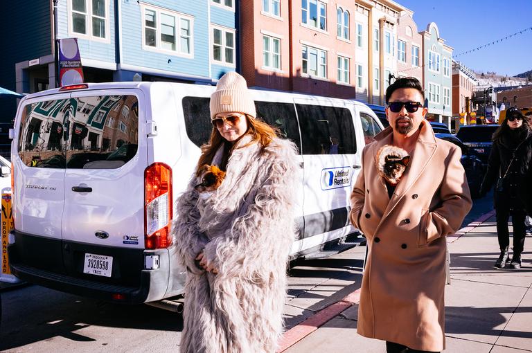 A couple walks past a parked van on Main Street, she in a lavender fur coat and knit beanie, he in a camel overcoat carrying a small dog.