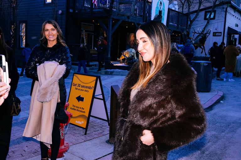 Two women walking past storefronts at dusk, one smiling in a black jacket with a pink scarf, the other in a dark fur coat. An Audible sign glows in the background.