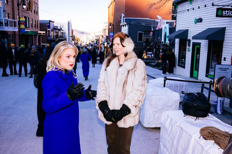 Two women standing on Main Street at golden hour, one in a royal blue coat with red lipstick, the other in a cream fur coat and black gloves.