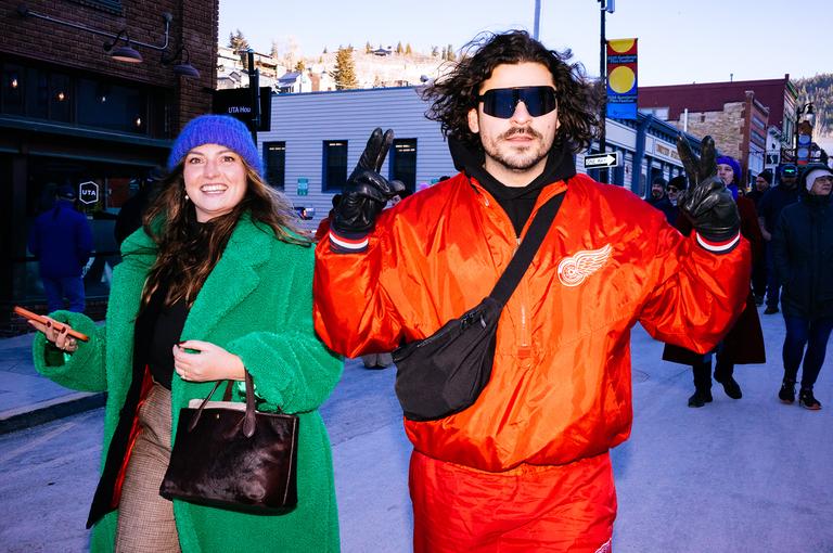 A woman in a green overcoat and blue beanie walks alongside a man in an oversized orange Detroit Red Wings jacket and blue sunglasses.