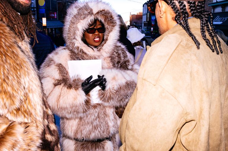 A woman in a fur-hooded coat and sunglasses stands among a group on a crowded sidewalk, framed tightly between fur coats.