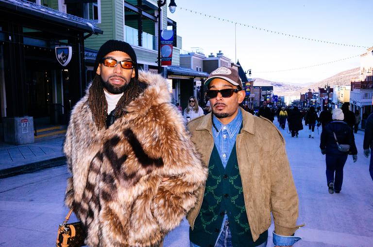 Two men in fur and suede coats with orange-tinted sunglasses walk confidently down Main Street, the snowy mountains visible in the distance.
