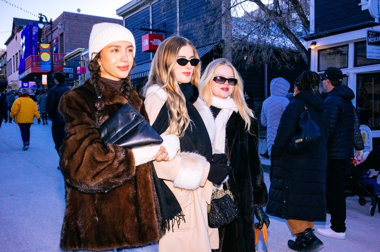 Three women pose together on Main Street at dusk, wearing fur coats and sunglasses, the festival crowd flowing behind them.