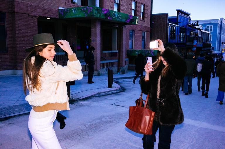 A woman adjusts her wide-brimmed hat while her friend photographs her with a phone near the Onyx building at blue hour.