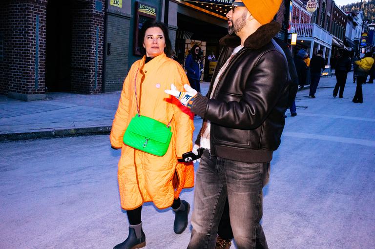 A couple walks down Main Street at blue hour, she in an oversized yellow quilted coat with a bright green crossbody bag, he in a leather jacket and orange beanie.