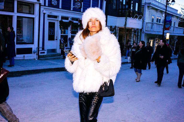 A woman in an all-white fur coat and fur hat walks alone down Main Street, phone in hand, the evening crowd blurred behind her.